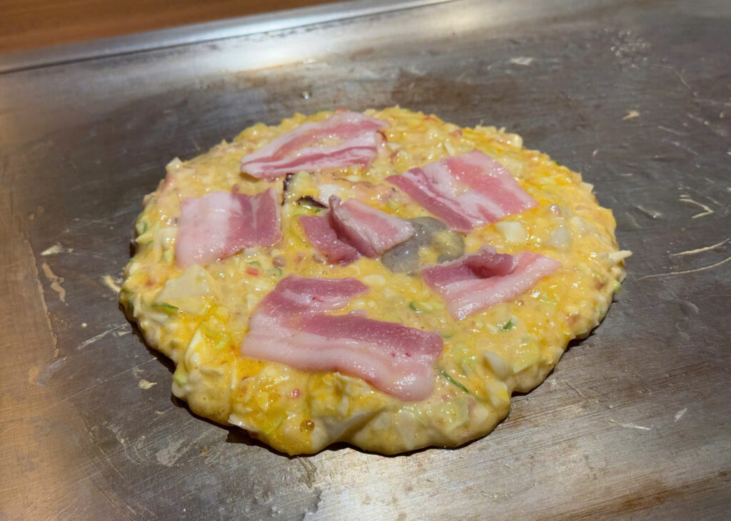 Chef preparing Osaka-style okonomiyaki with cabbage and pork on a hot iron griddle